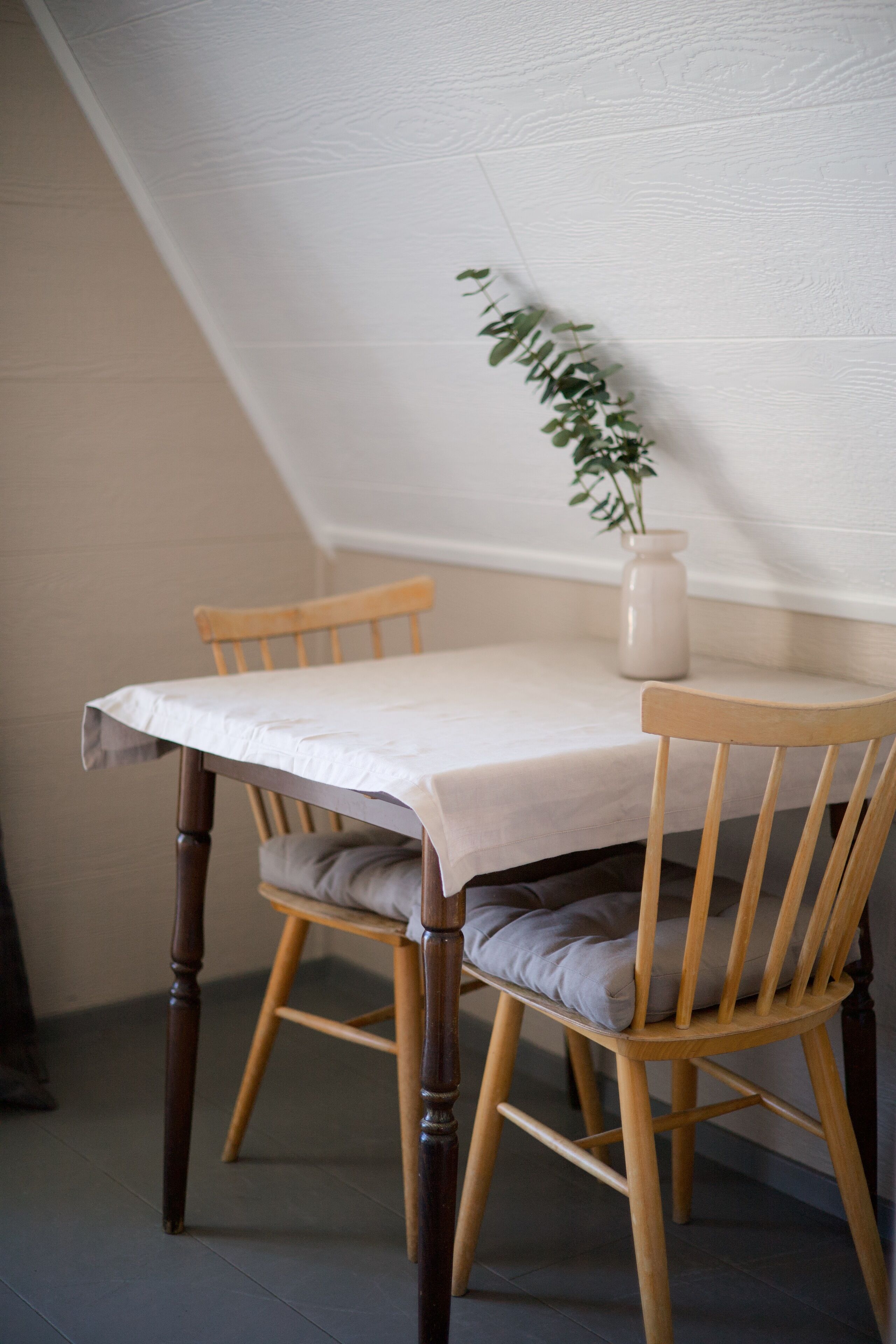 A dining table with a white tablecloth, two wooden chairs, a cushion on one chair, and a vase with a plant on the table.