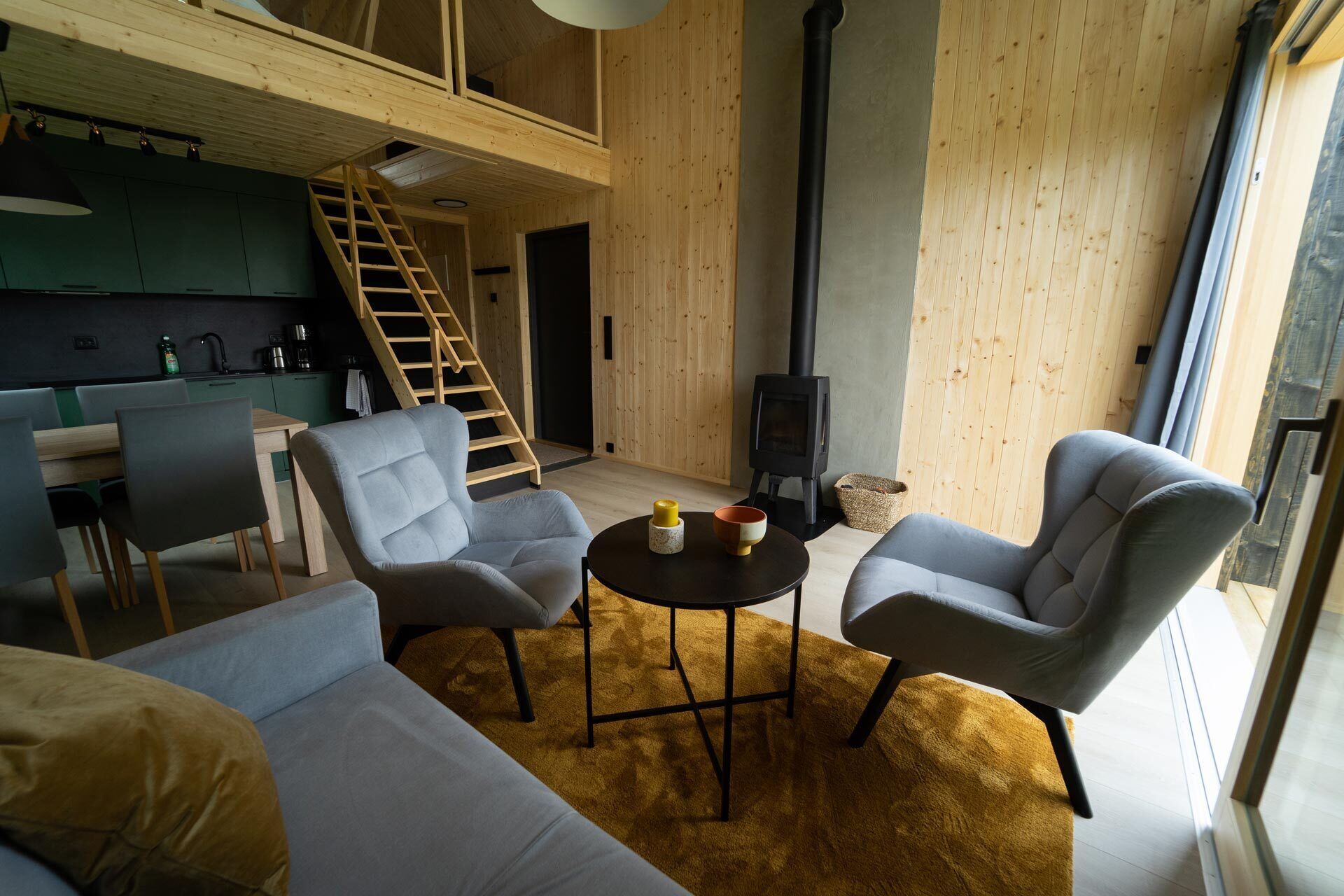 A modern living room with a high ceiling, a staircase, a kitchen area, and a fireplace.