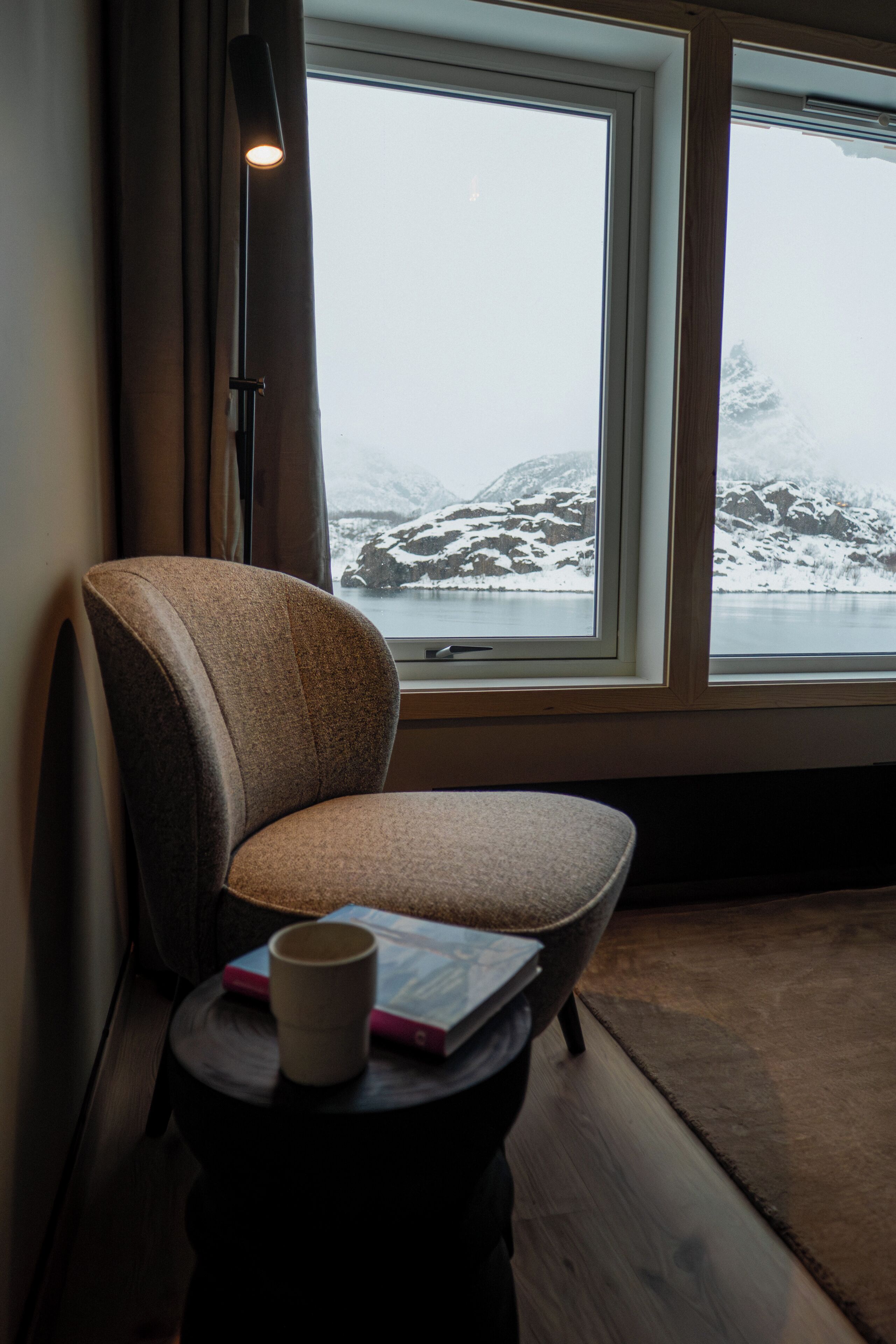 A room with a window, a chair, a small table with a book and a mug.
