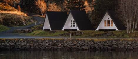Three white houses with black roofs by a lake, surrounded by trees and a stone wall.