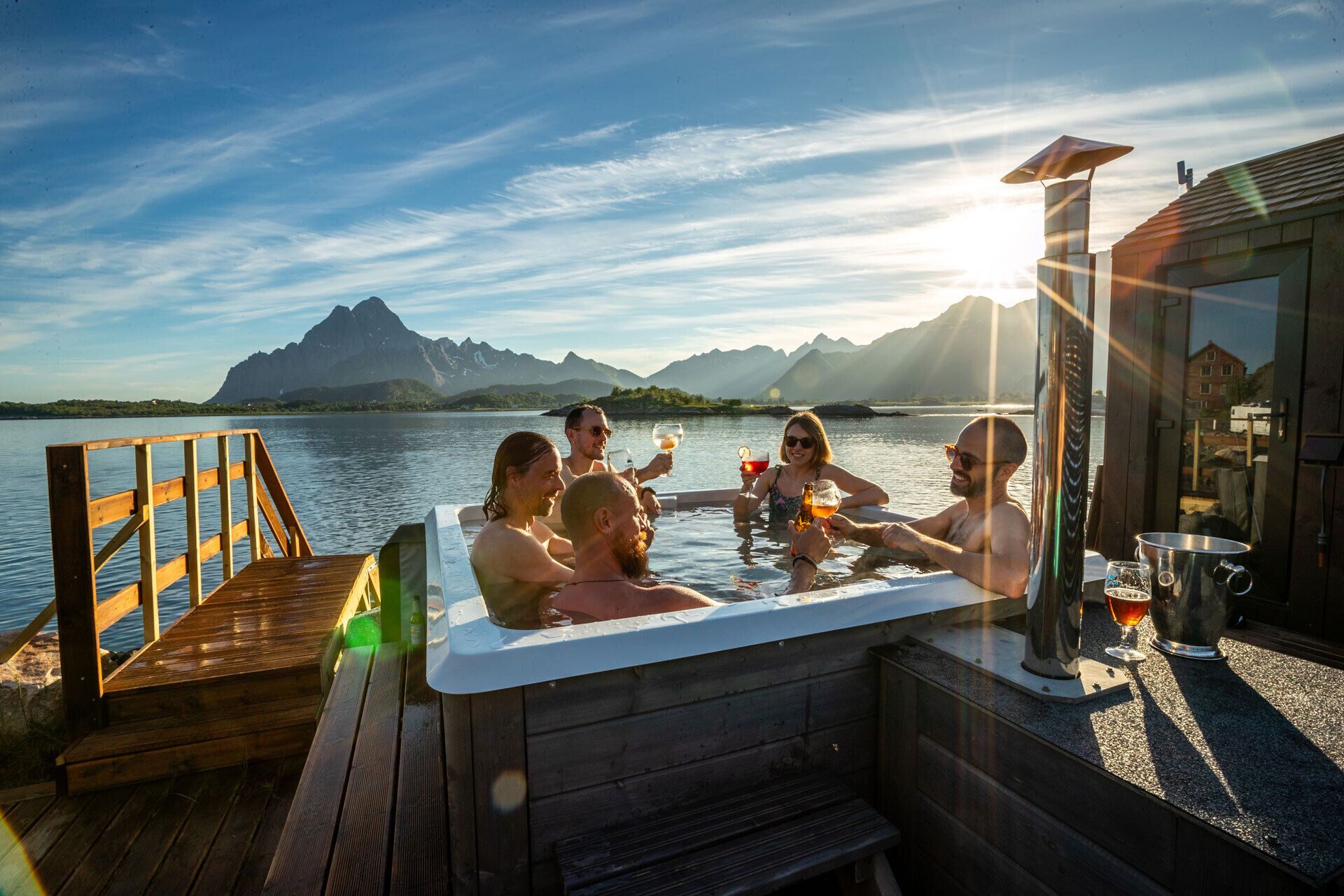 A group of people enjoying a drink in a hot tub on a wooden deck with a view of mountains and a body of water.