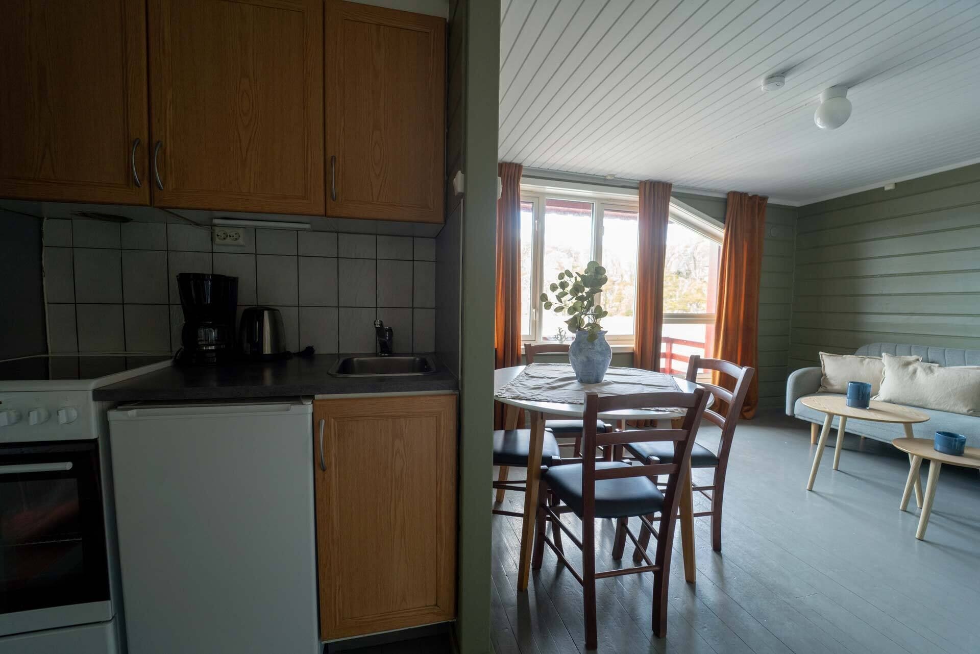 Une cuisine avec des meubles en bois, une machine à café et un évier. On y trouve également un coin repas avec une table, des chaises et un vase avec une plante. Un canapé et une petite table avec une tasse sont également visibles.