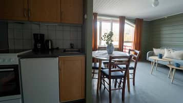 Cocina con gabinetes de madera, una cafetera y un fregadero. Hay un comedor con una mesa, sillas y un jarrón con una planta. También se ve un sofá y una pequeña mesa con una taza.
