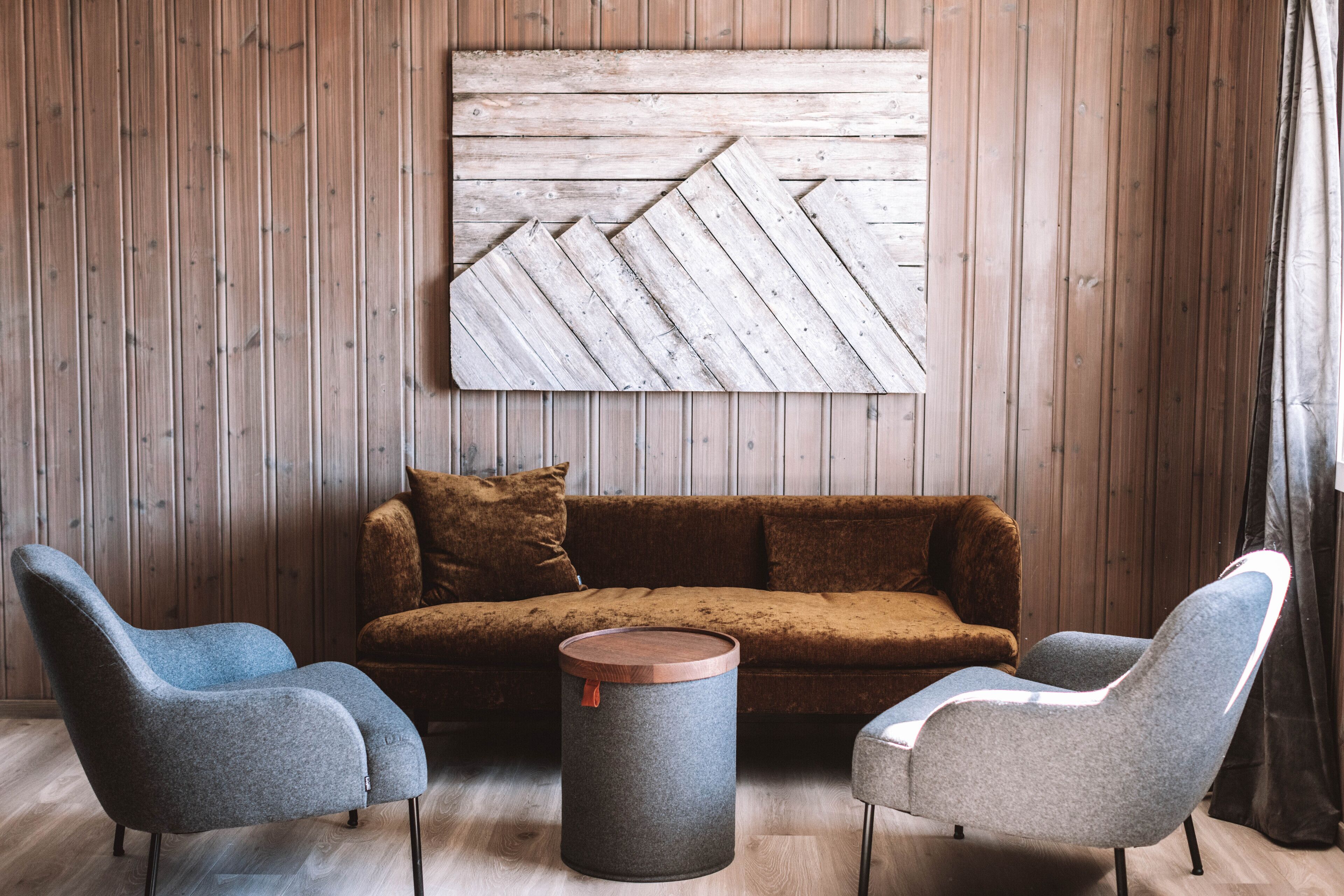 A room with a wooden wall, a brown sofa, two grey armchairs, and a round table with a wooden top.