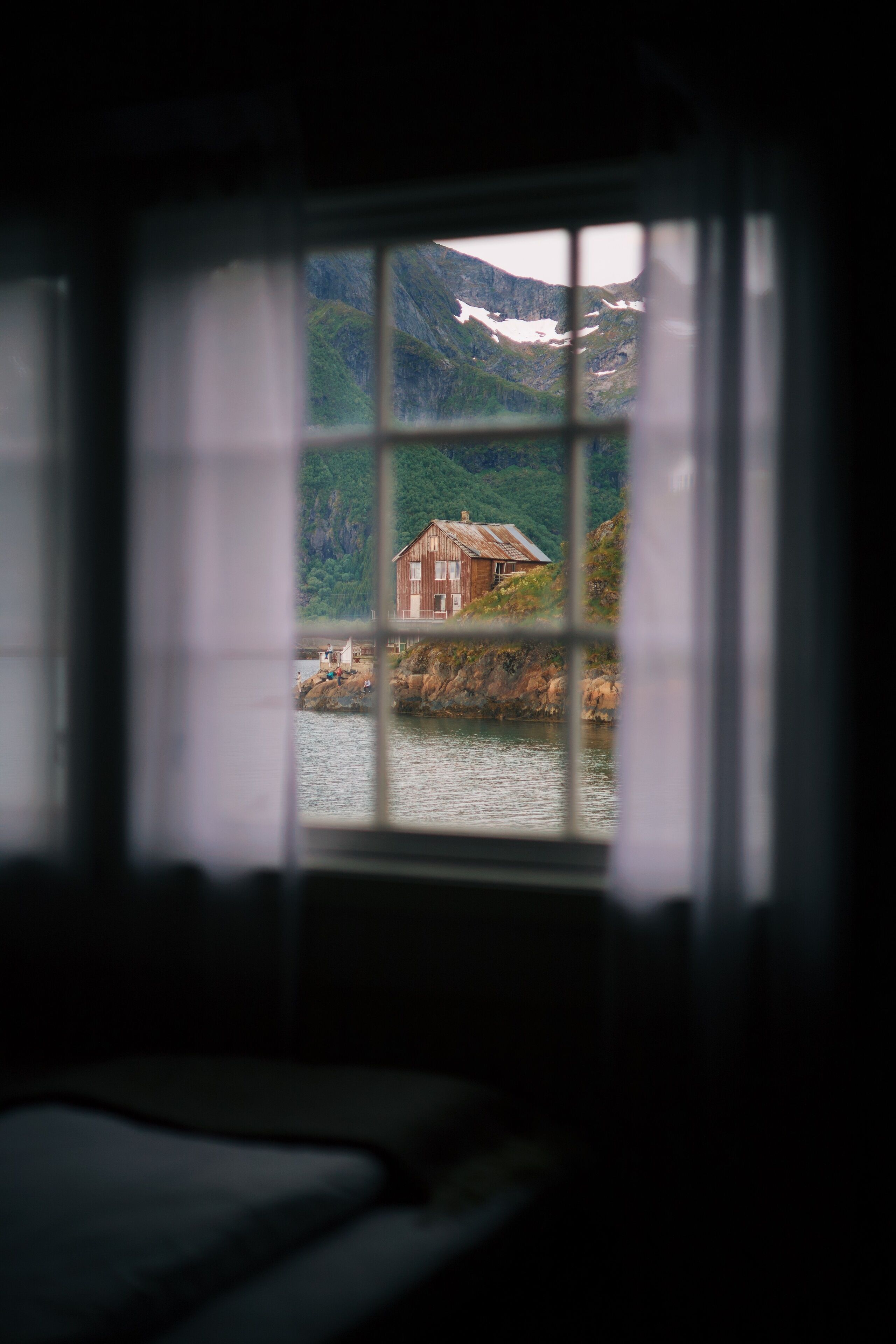 A room with a window overlooking a house by a body of water, surrounded by greenery and mountains.