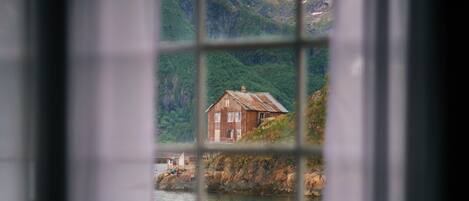 A room with a window overlooking a house by a body of water, surrounded by greenery and mountains.