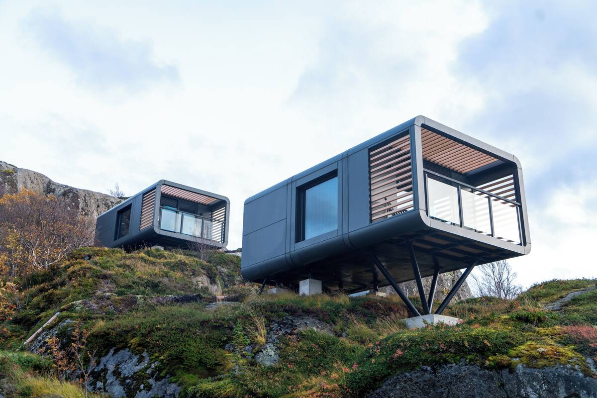 Two modern cabins with large windows, elevated on stilts, set on a rocky hillside with sparse vegetation.