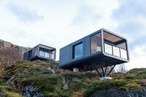 Dos cabañas modernas con ventanas amplias, elevadas sobre pilotes, ubicadas en una ladera rocosa con vegetación escasa.