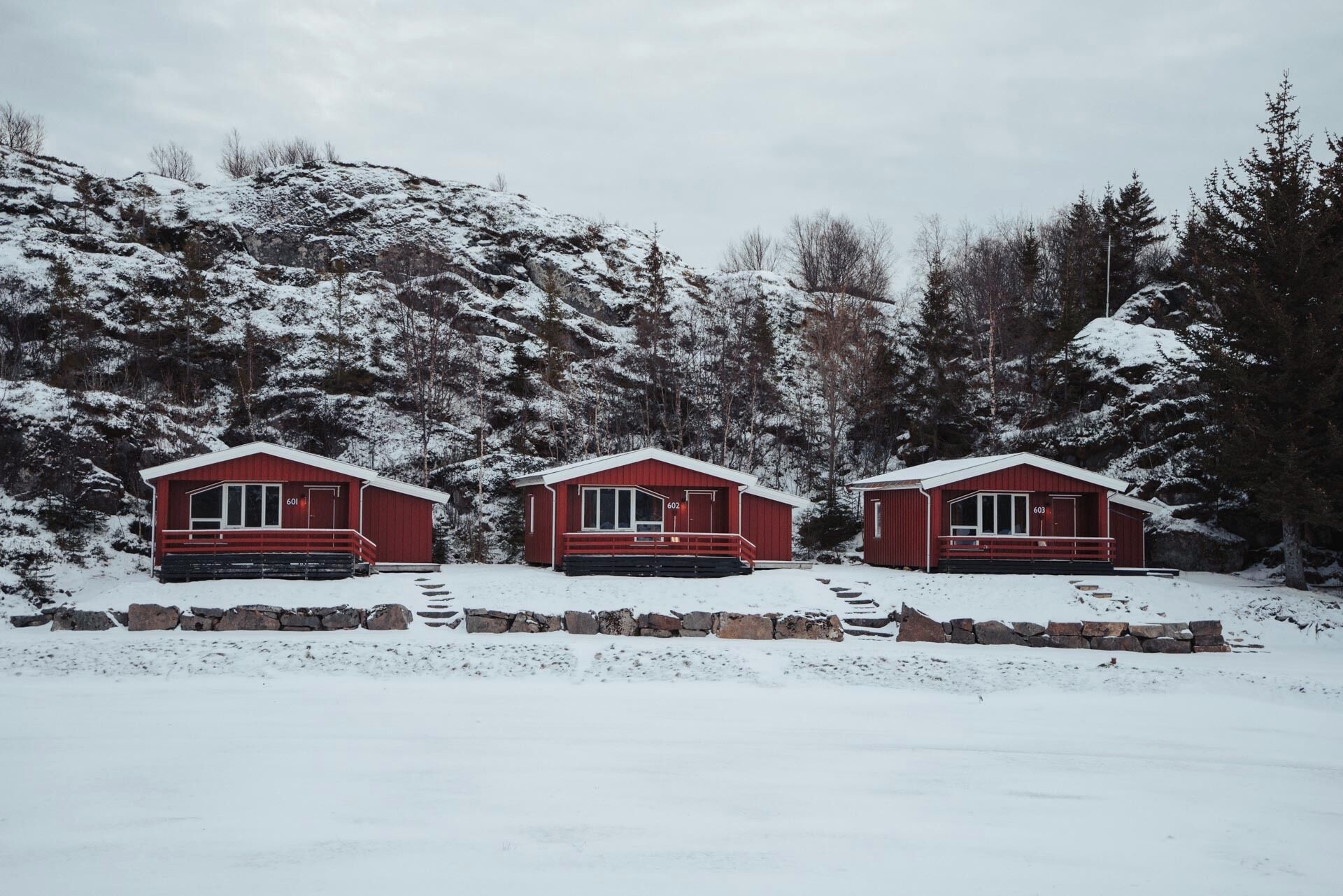 Three red cabins with numbered doors, set against a snowy backdrop with trees and a stone wall in the foreground.