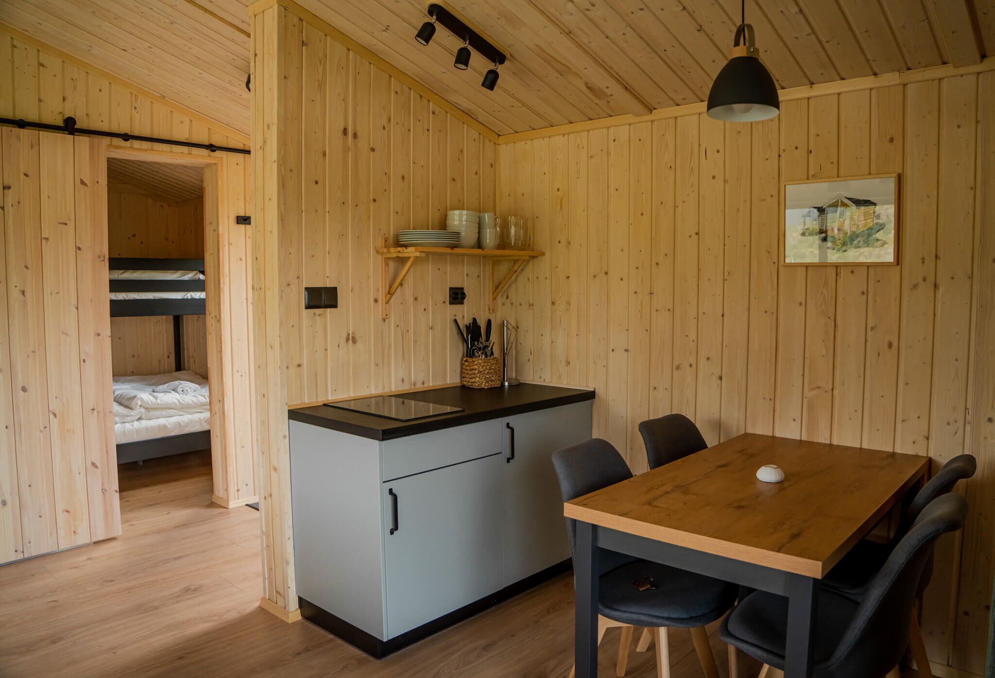 A compact room with a kitchenette, a dining table with chairs, a bunk bed, and wooden walls.