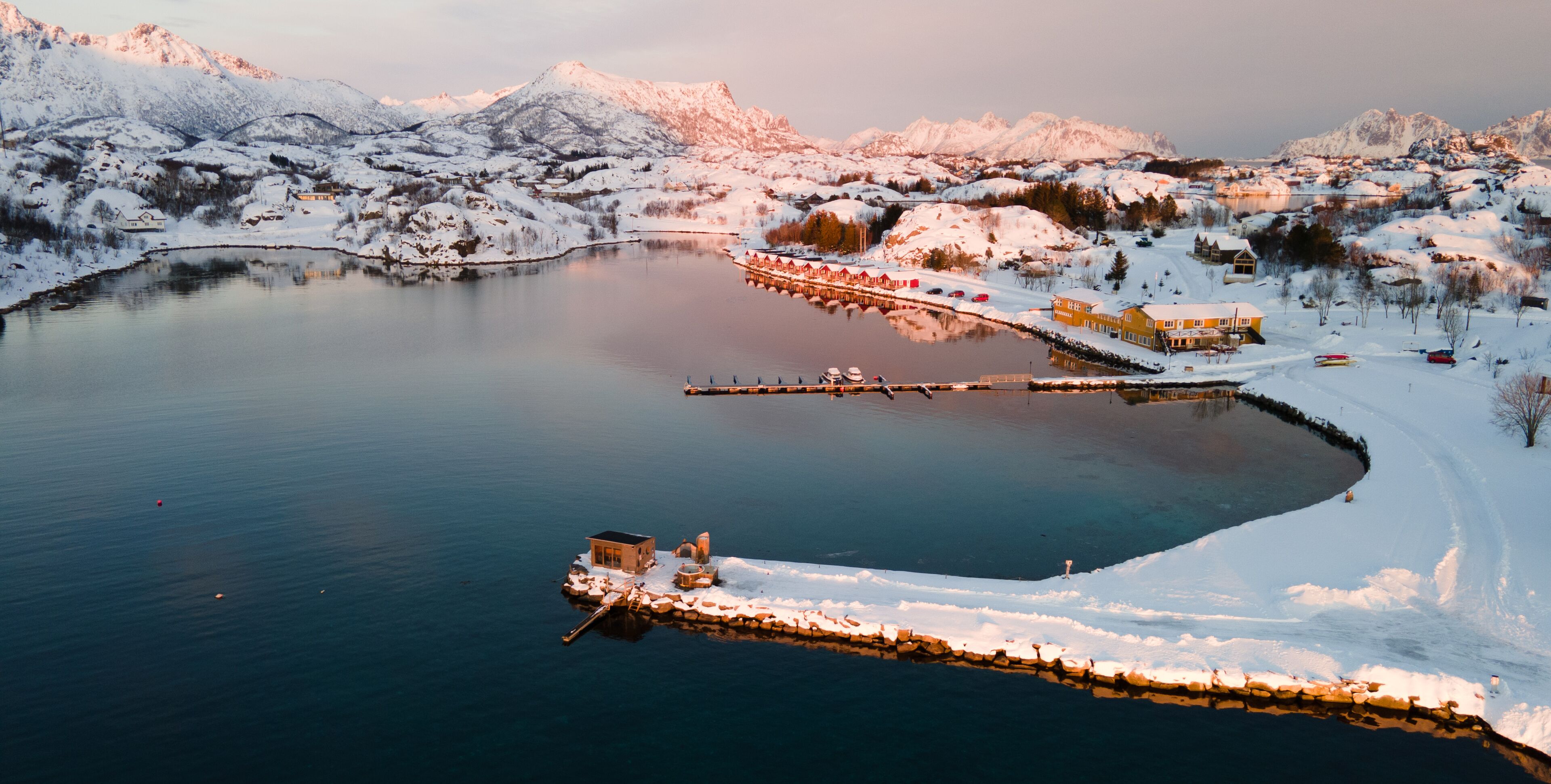 Een besneeuwd dorp aan een meer met een steiger die in het water reikt, omringd door bergen.