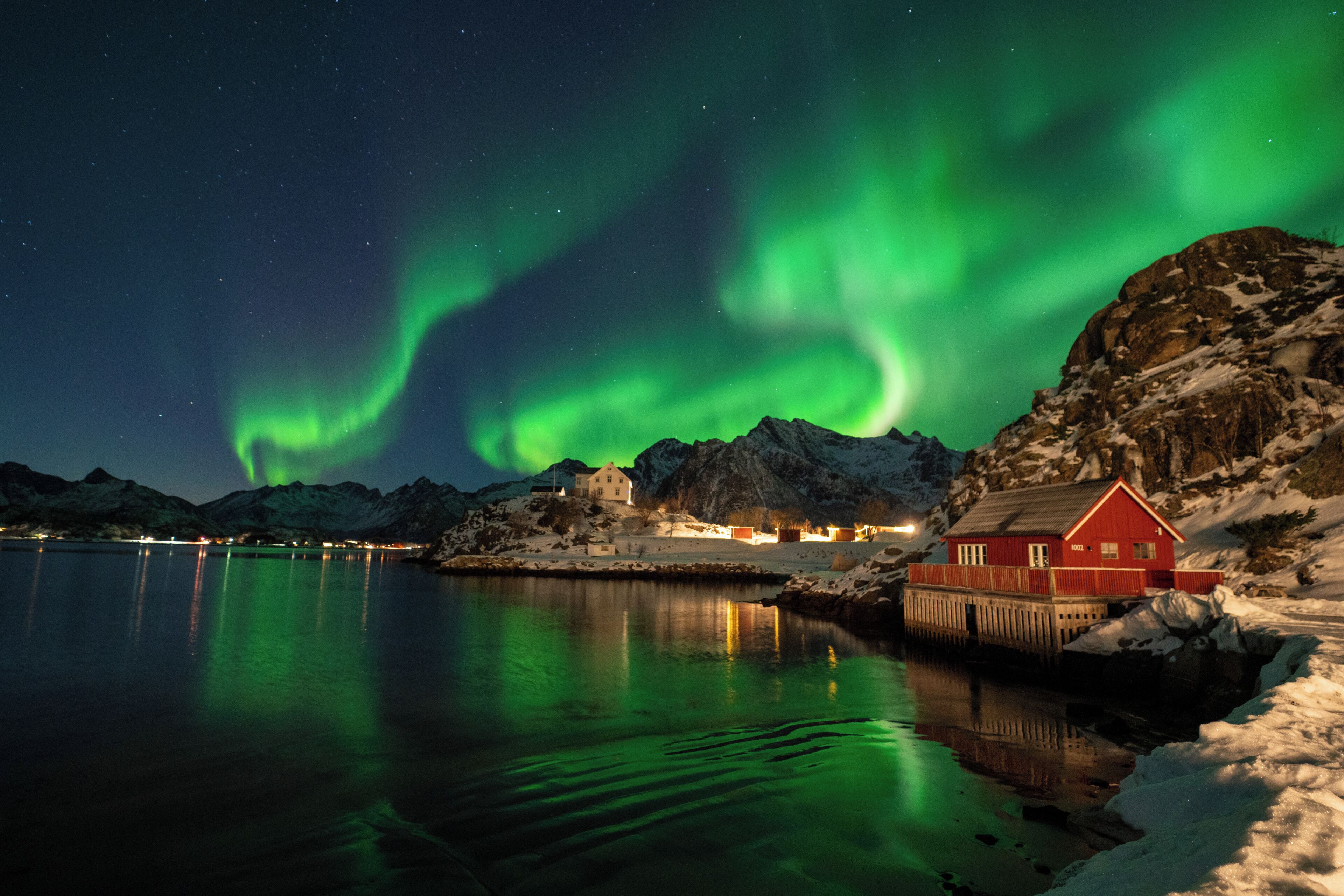 Paesaggio costiero con un edificio rosso vicino a una superficie d'acqua tranquilla, che riflette luci e il cielo notturno.