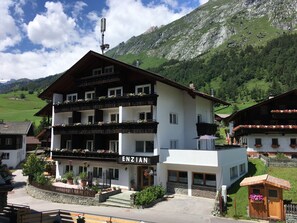 A multi-story hotel with balconies adorned with flowers, a sign reading "ENZIAN," and a mountainous backdrop. - Gästehaus Enzian (Praegraten am Grossvenediger)