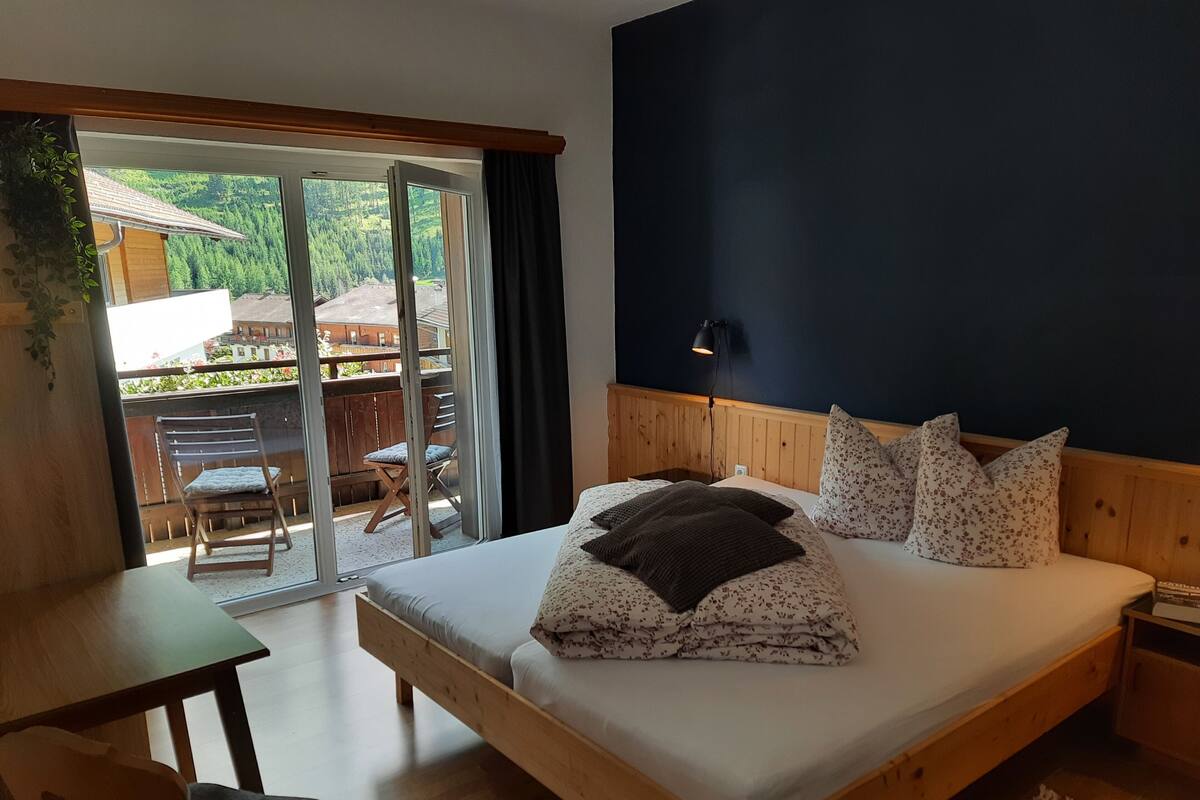 Ein Schlafzimmer mit einem Holzbett, einem Schreibtisch und einem Stuhl. Es bietet einen Blick auf einen Balkon mit Holzzaun und einem Gebäude im Hintergrund.