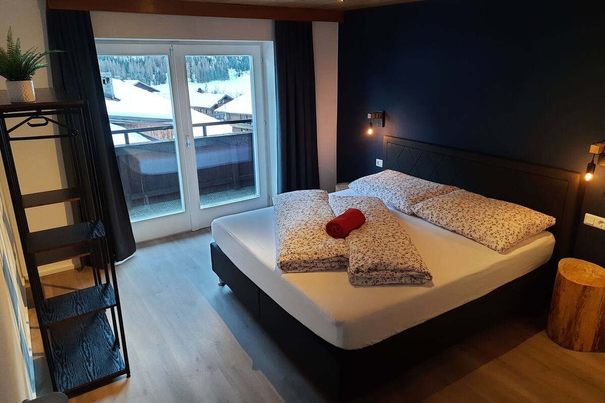 Ein Schlafzimmer mit einem großen Bett, einem Holz-Kopfteil, einem Nachttisch, Holzboden, einem Fenster mit Blick auf eine verschneite Landschaft und einer Pflanze auf einem Regal.
