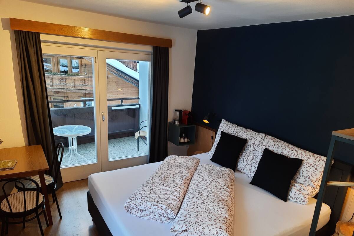 Ein Hotelzimmer mit Bett, Tisch, Stühlen, einem kleinen Balkon mit Tisch und Stühlen sowie Blick auf ein Gebäude.