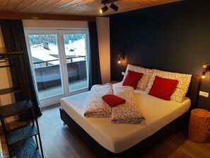 A bedroom with a large bed, a wooden headboard, a wooden floor, a sliding glass door, and a view of a snowy landscape. - Gästehaus Enzian (Praegraten am Grossvenediger)