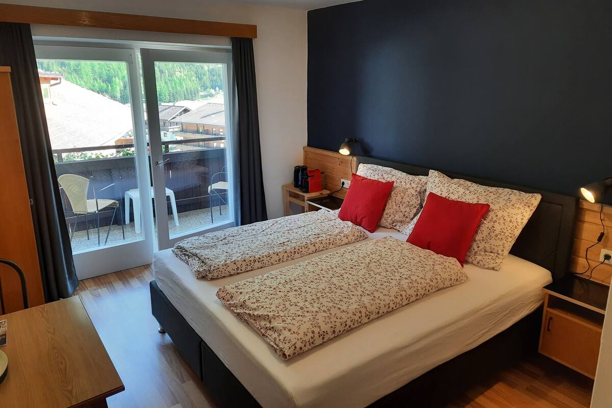 Ein Schlafzimmer mit einem großen Bett, einem Nachttisch, einem Holzkopfende, einem Fenster mit Vorhängen und Blick ins Freie.