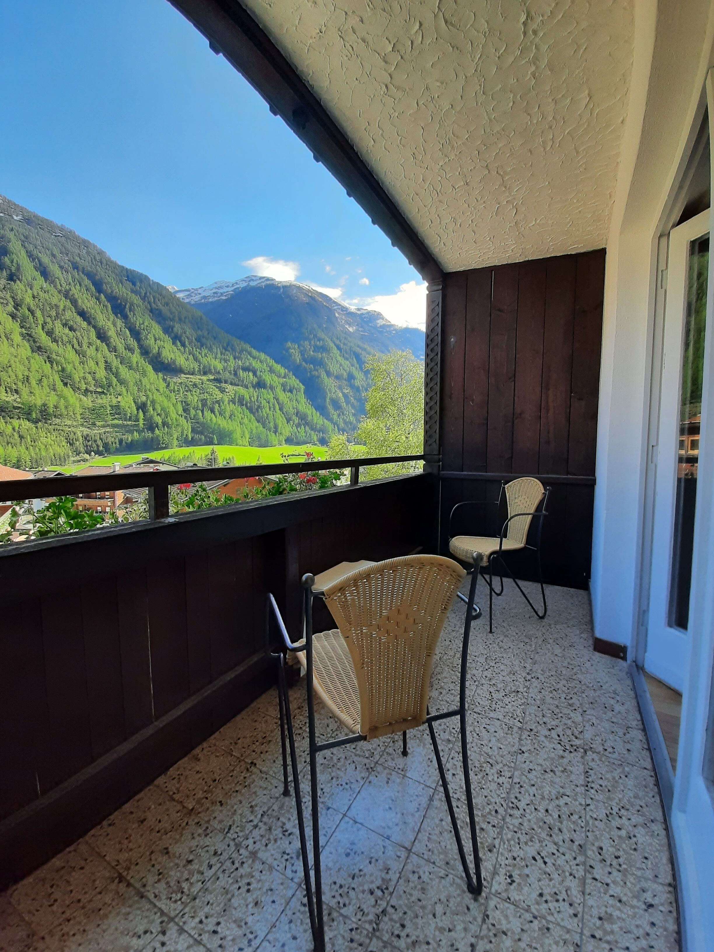 Un balcon avec deux fauteuils en osier, un sol carrelé et une vue sur des montagnes verdoyantes.