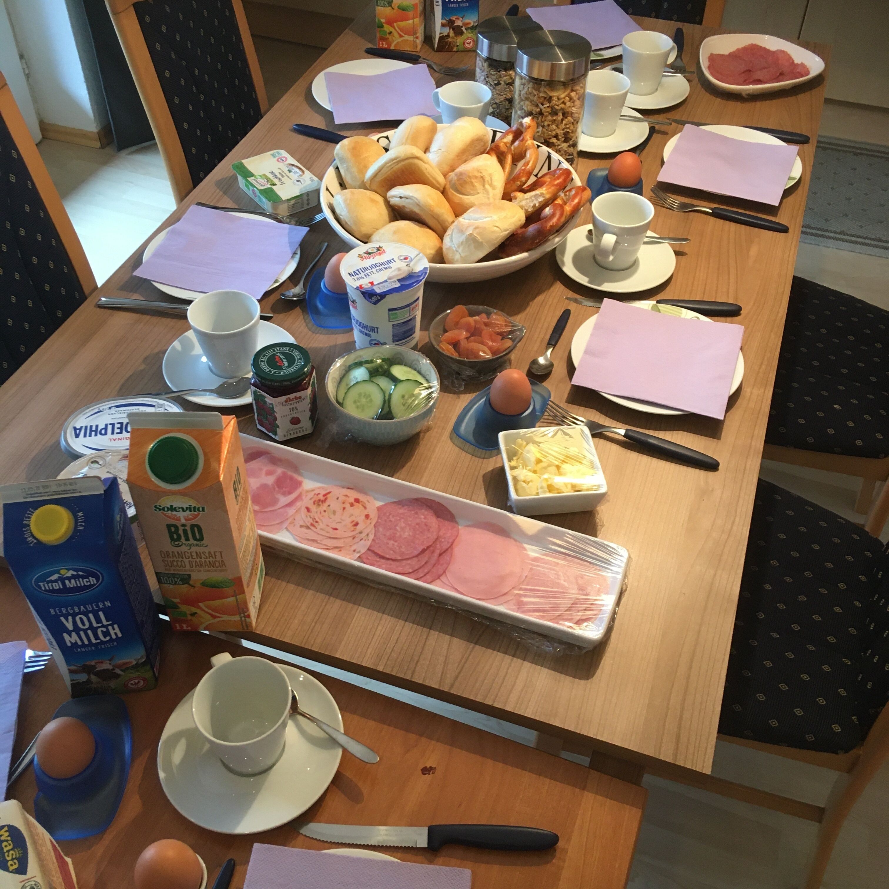 A breakfast table with various food items, including bread, sausages, eggs, and a tray of sliced meat.
