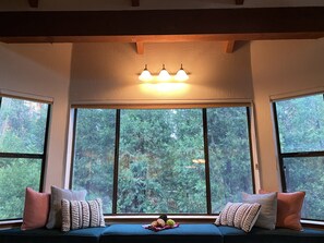 A cozy seating area with a view of a forest through large windows, a ceiling light fixture, and a small table with a fruit bowl. - Beautiful Cedar Cabin (Northfork)