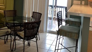 A dining area with a glass table and wicker chairs, a kitchen with a white countertop, and a sliding glass door leading to a balcony. - SANDY TAILS - Oak Island, NC (Oak Island)
