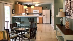 A compact kitchen with wooden cabinets, a white refrigerator, a microwave, and a dining area with wicker chairs and a glass table. - SANDY TAILS - Oak Island, NC (Oak Island)