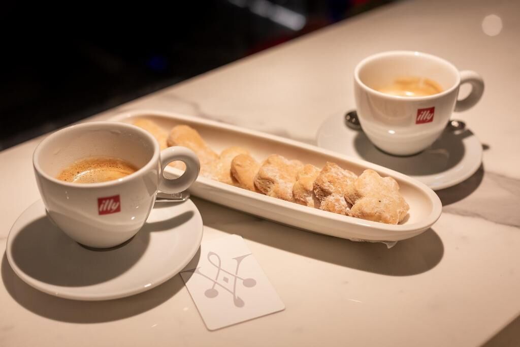 A table with two white cups of coffee and a plate of pastries.