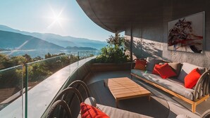 Un balcon aménagé avec un canapé, une table basse et des chaises, offrant une vue panoramique sur les montagnes et un plan d’eau.