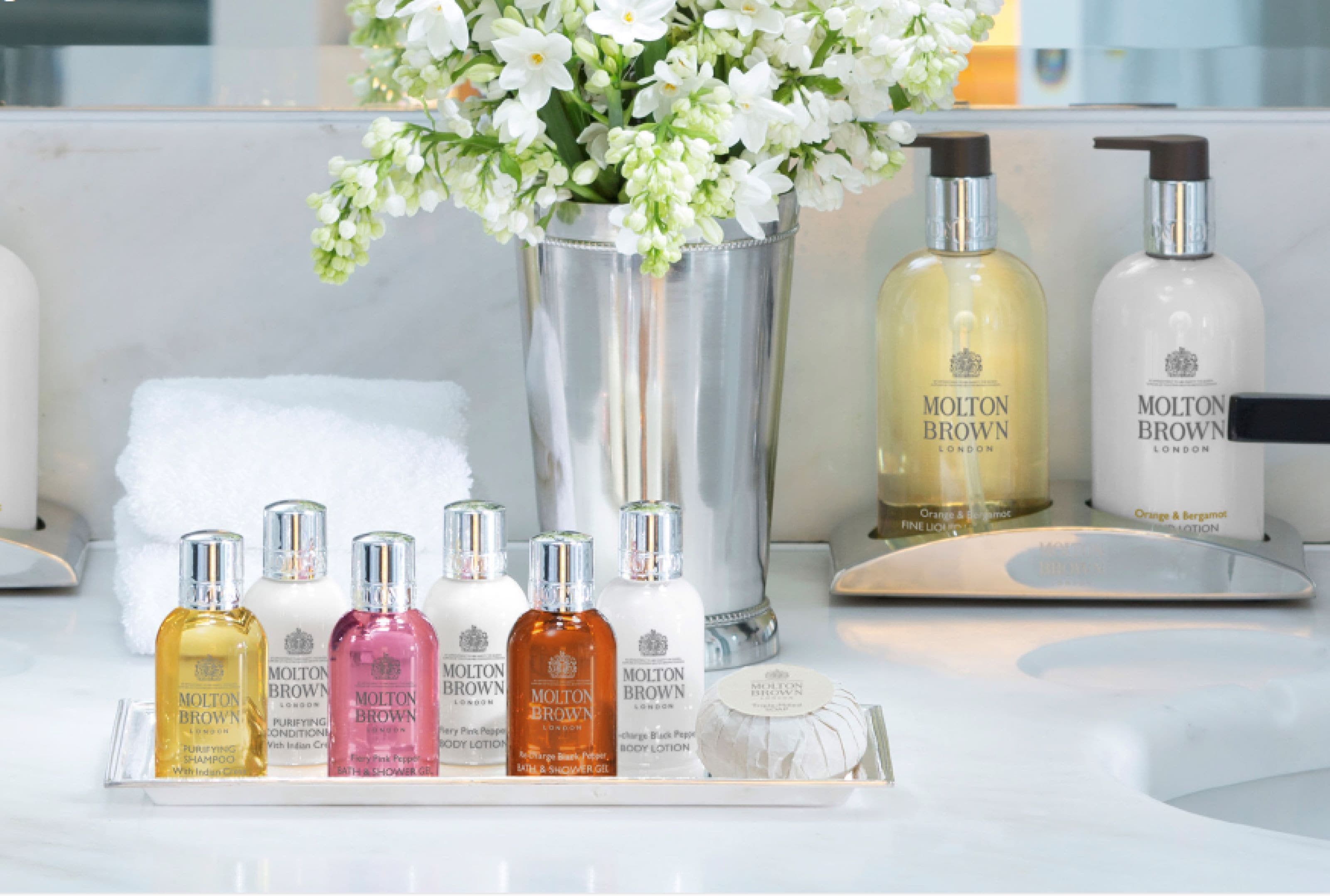 Un tocador de baño con una bandeja que contiene varios artículos de tocador Molton Brown, un jarrón con flores blancas y una toalla blanca.
