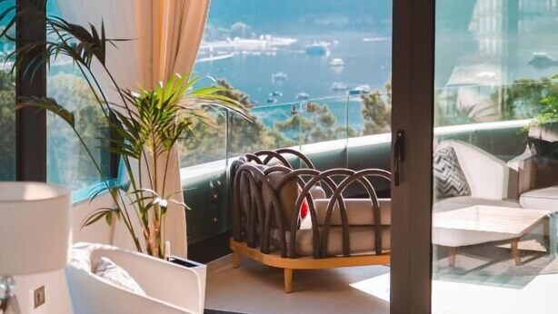 Un salon moderne doté d’une grande fenêtre offrant une vue imprenable sur la mer et les bateaux, d’un lustre et d’un coin salon confortable.
