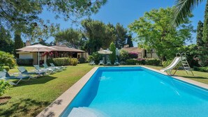 Una piscina con sillas de descanso y un tobogán, rodeada de árboles y un edificio con tejado de baldosas.