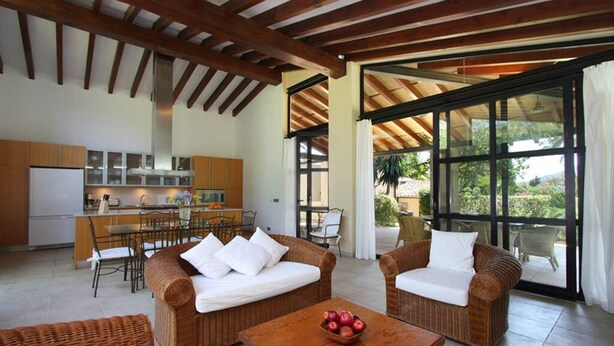 Un salón amplio con muebles de mimbre, una mesa de madera con un frutero, y una cocina al fondo.