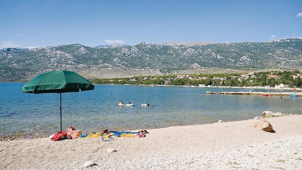 Une plage avec un parasol vert, des gens qui prennent un bain de soleil et une montagne en arrière-plan.