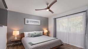 Une chambre à coucher avec un lit, deux lampes de chevet, un ventilateur de plafond, une fenêtre avec des rideaux légers et un tableau encadré accroché au mur.