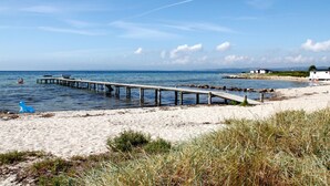 Une plage avec une jetée en bois s’avançant dans des eaux calmes, une petite sculpture bleue et une maison située au loin.