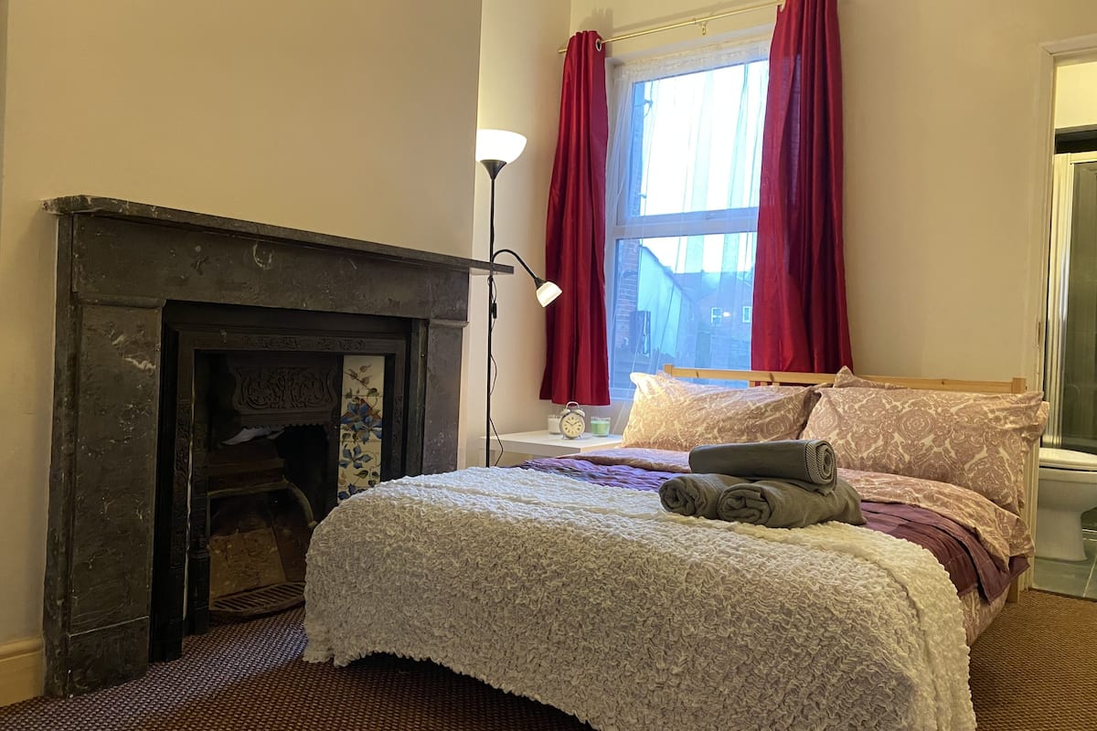 A bedroom with a fireplace, a bed with patterned bedding, a nightstand, and a window with red curtains.