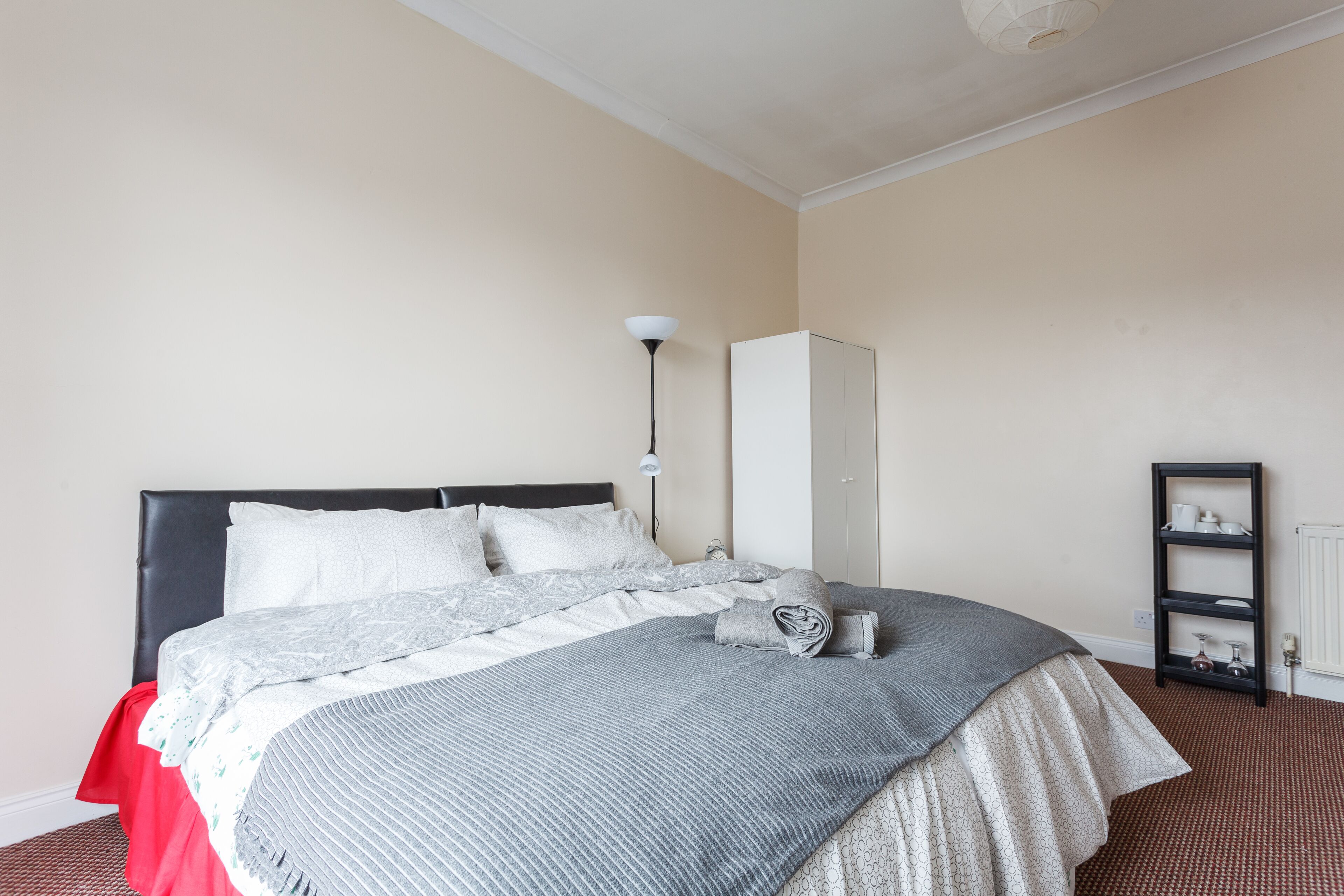 Ein ordentlich bezogenes Bett mit einer grau-weißen Decke, einem schwarzen Kopfteil und einem roten Fußteil. An der Wand befinden sich ein weißer Kleiderschrank und ein schwarzes Regalsystem.