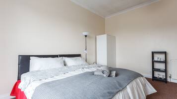 Ein ordentlich bezogenes Bett mit einer grau-weißen Decke, einem schwarzen Kopfteil und einem roten Fußteil. An der Wand befinden sich ein weißer Kleiderschrank und ein schwarzes Regalsystem.