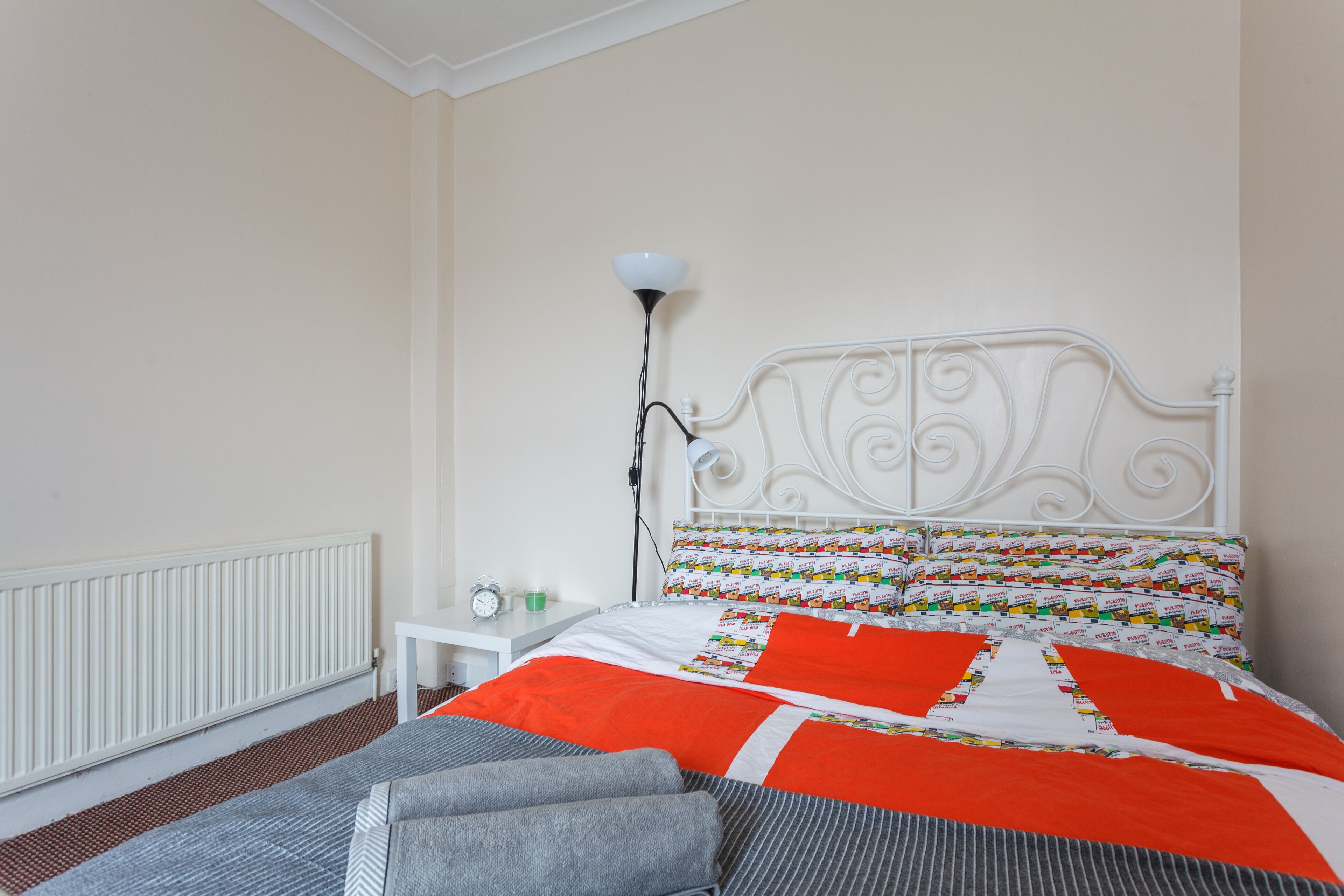 A neatly made bed with a patterned quilt, a white bedside table with a lamp, and a radiator on the wall.