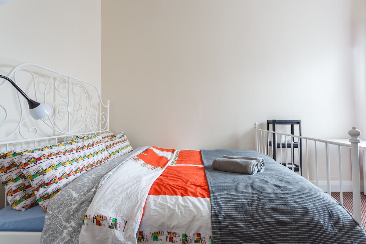 A neatly made bed with a white and orange quilt, a grey blanket, and a rolled grey towel.