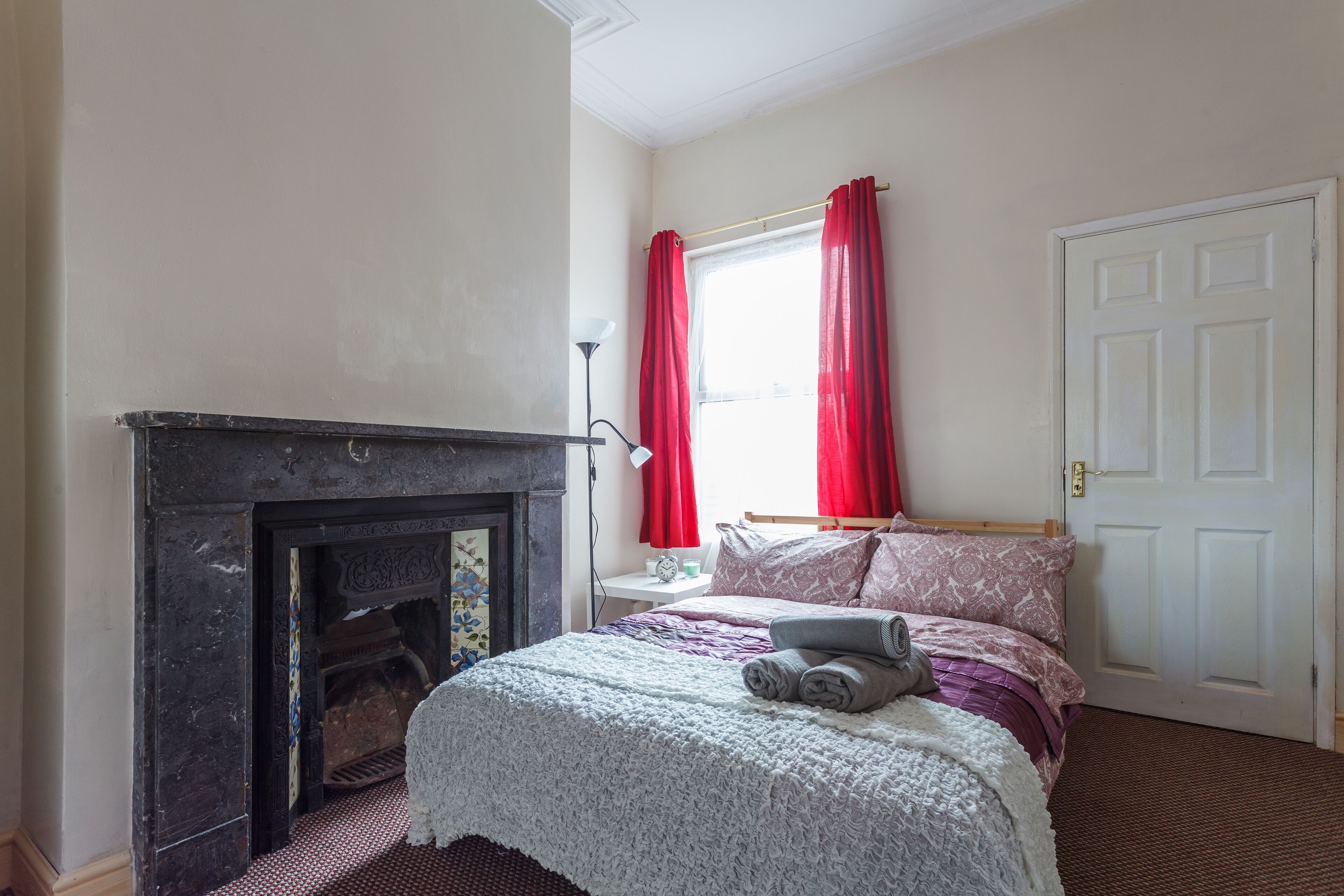 Quarto com lareira, cama com roupa de cama rosa, janela com cortinas vermelhas e porta branca.