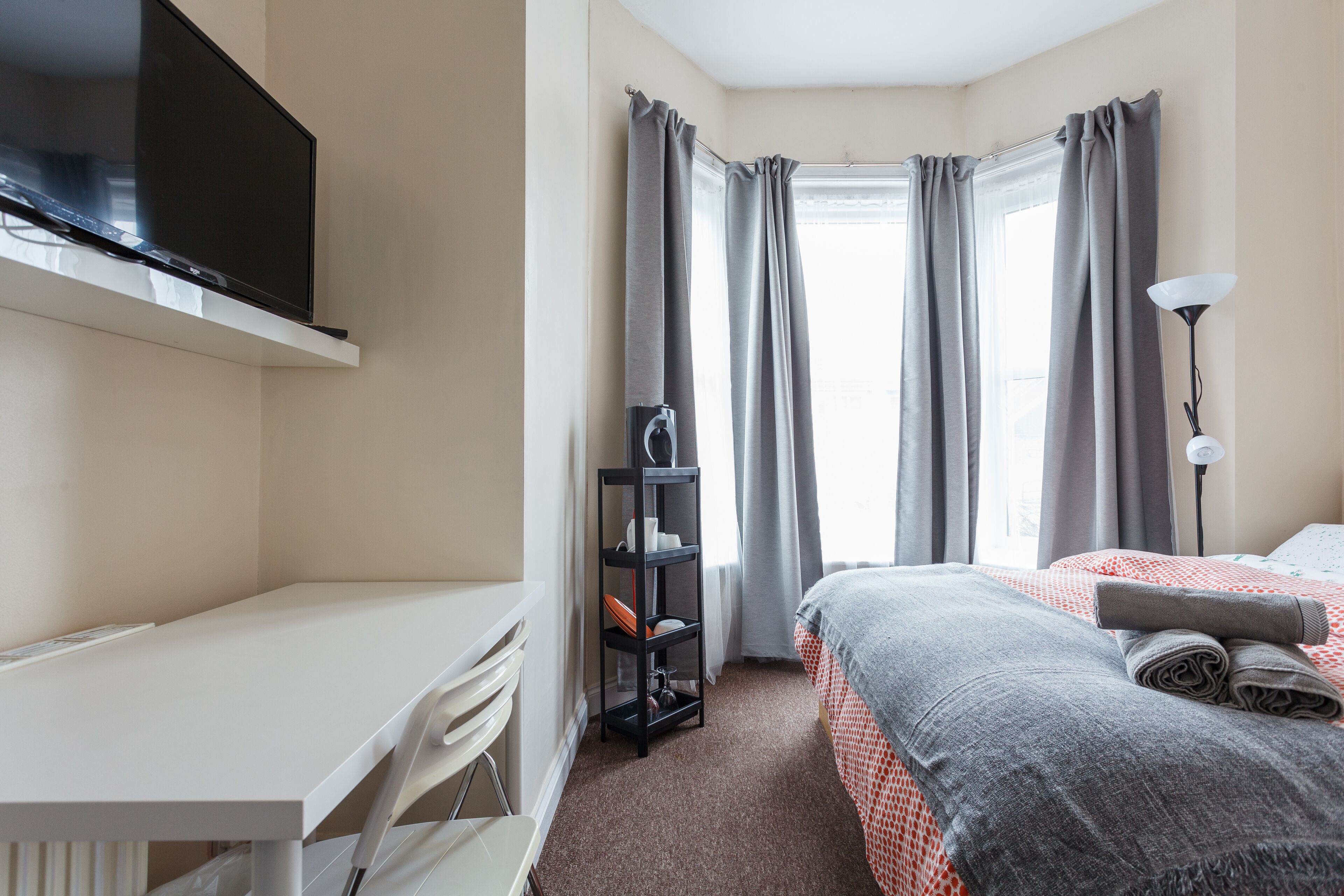 A bedroom with a bed, a desk, a TV, a shelf, and a window with curtains.
