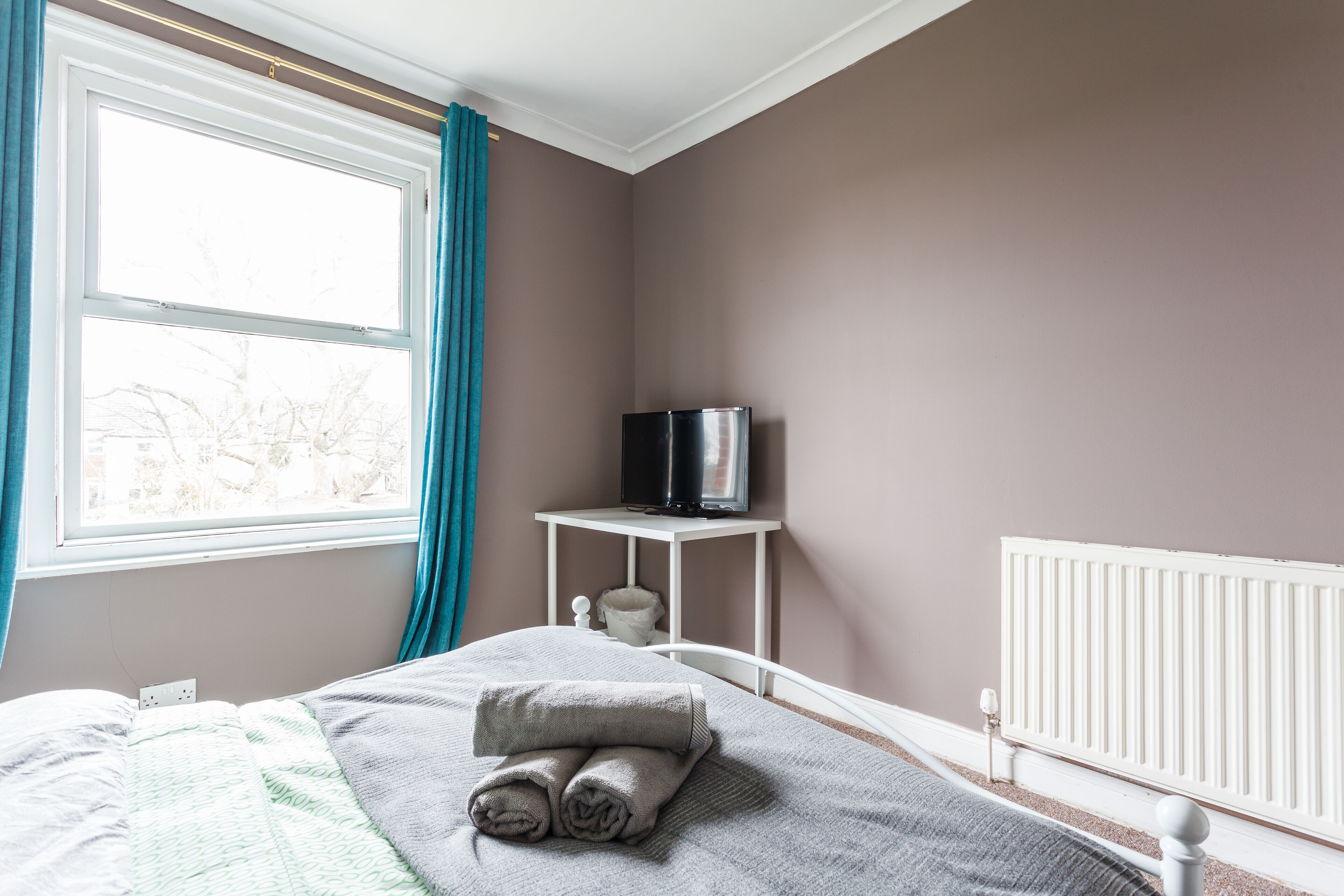 A bedroom with a bed, a TV on a small table, a radiator, and a window with curtains.