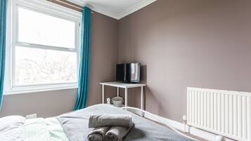 A bedroom with a bed, a TV on a small table, a radiator, and a window with curtains.