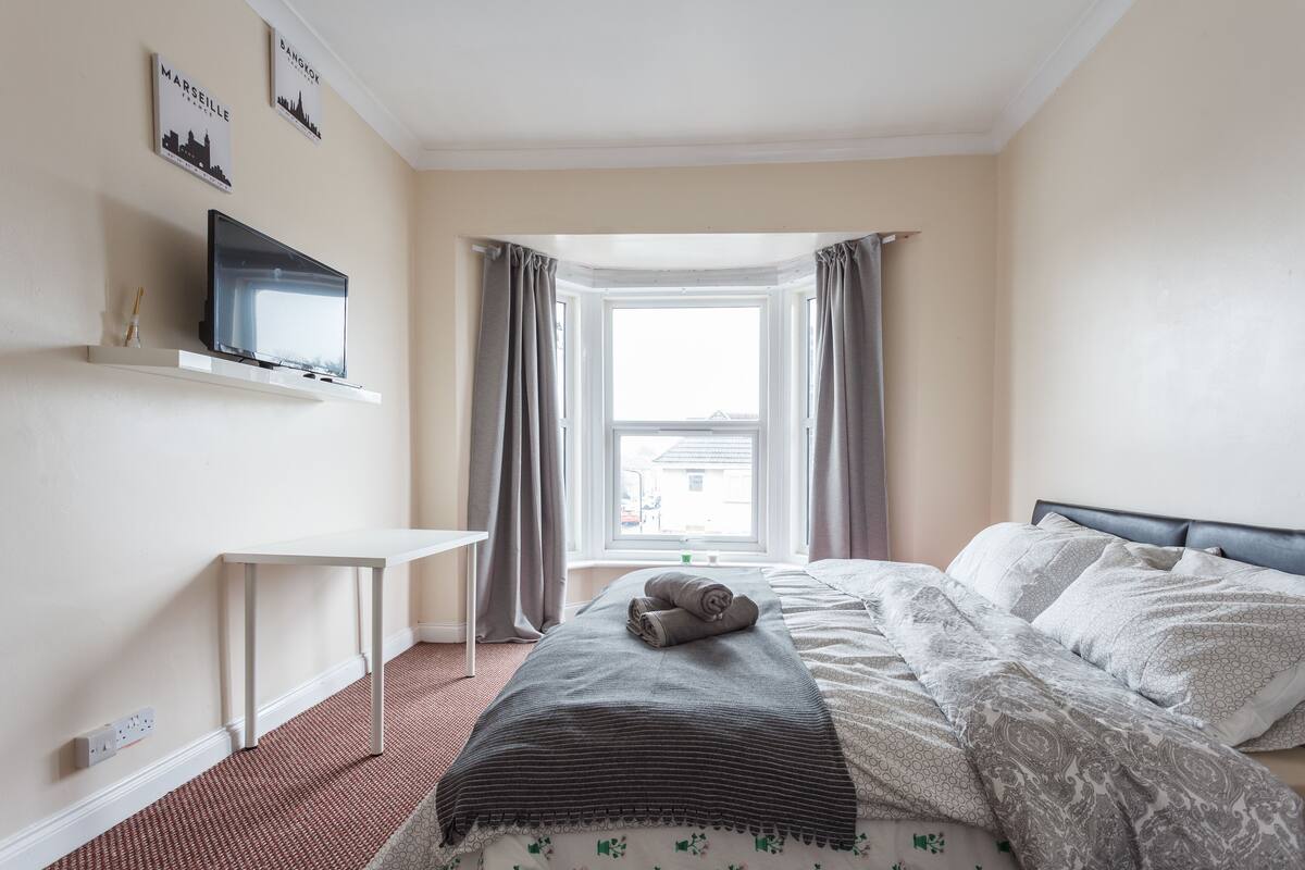 A bedroom with a bed, a TV on a shelf, a window with curtains, and a small table.