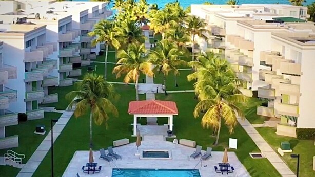 A resort with a central swimming pool, surrounded by lounge chairs and palm trees.