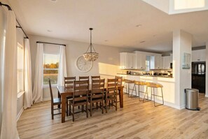 Une cuisine moderne avec des meubles blancs, un îlot central et un coin repas agrémenté de chaises en bois et d’un lustre.