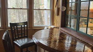 A wooden interior with a round table and two chairs, large windows with a view of trees, and a window blind. - Waterfront Camp with Boathouse - Wifi, kayaks, canoe and sandy bottom out front. (Old Forge)