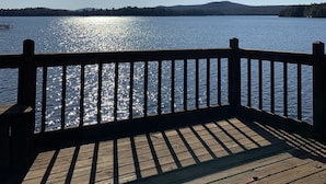 A wooden deck with a view of a calm lake and distant hills. - Waterfront Camp with Boathouse - Wifi, kayaks, canoe and sandy bottom out front. (Old Forge)